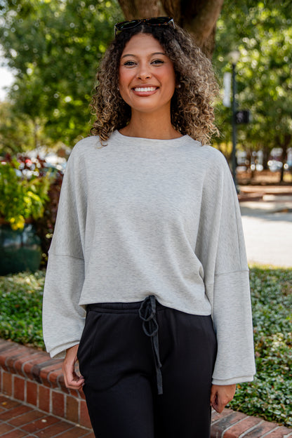 Woman wearing a gray sweater and black pants standing outdoors with greenery in the background