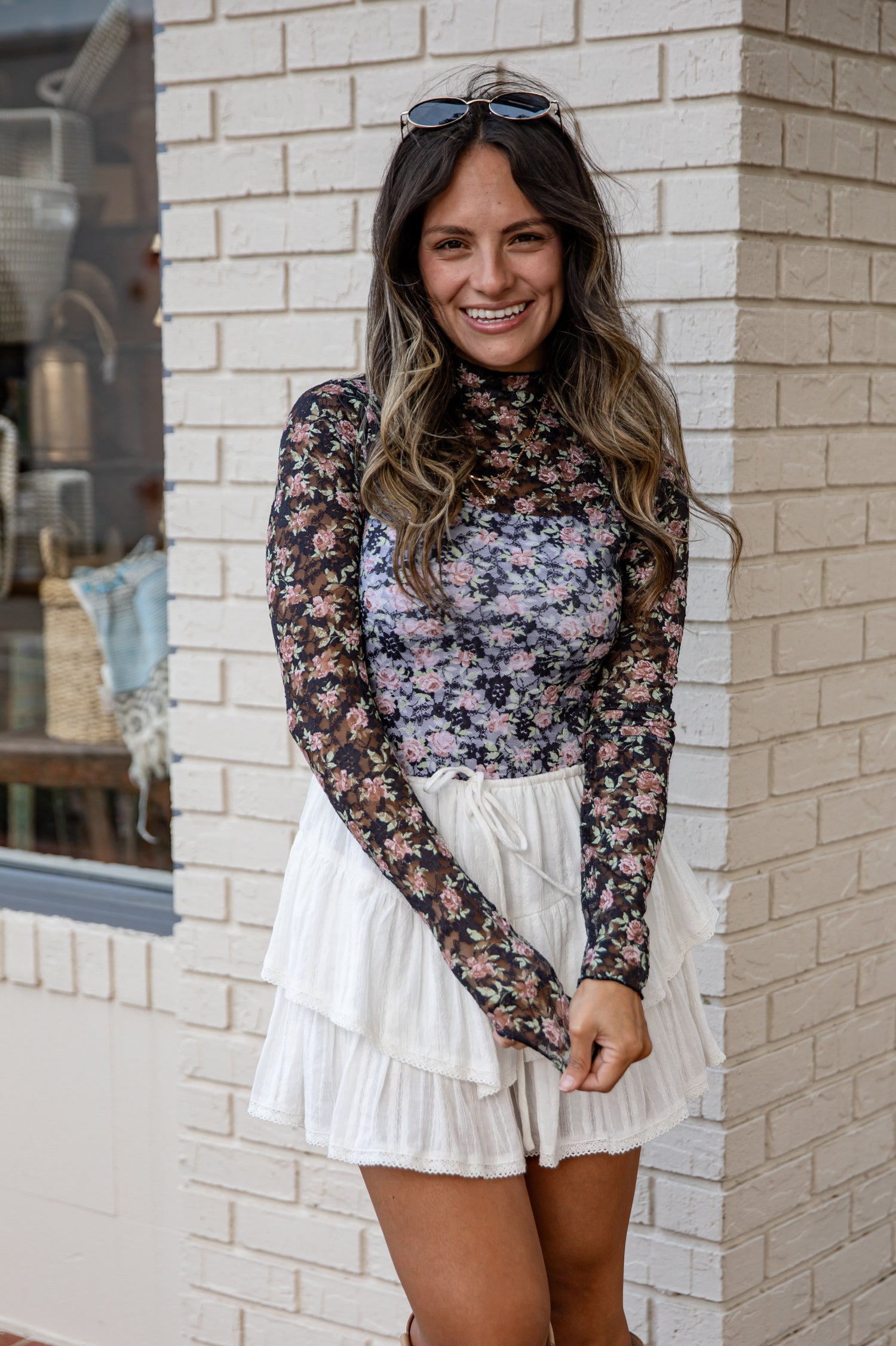 Woman wearing a floral long-sleeve top and white skirt standing against a brick wall.