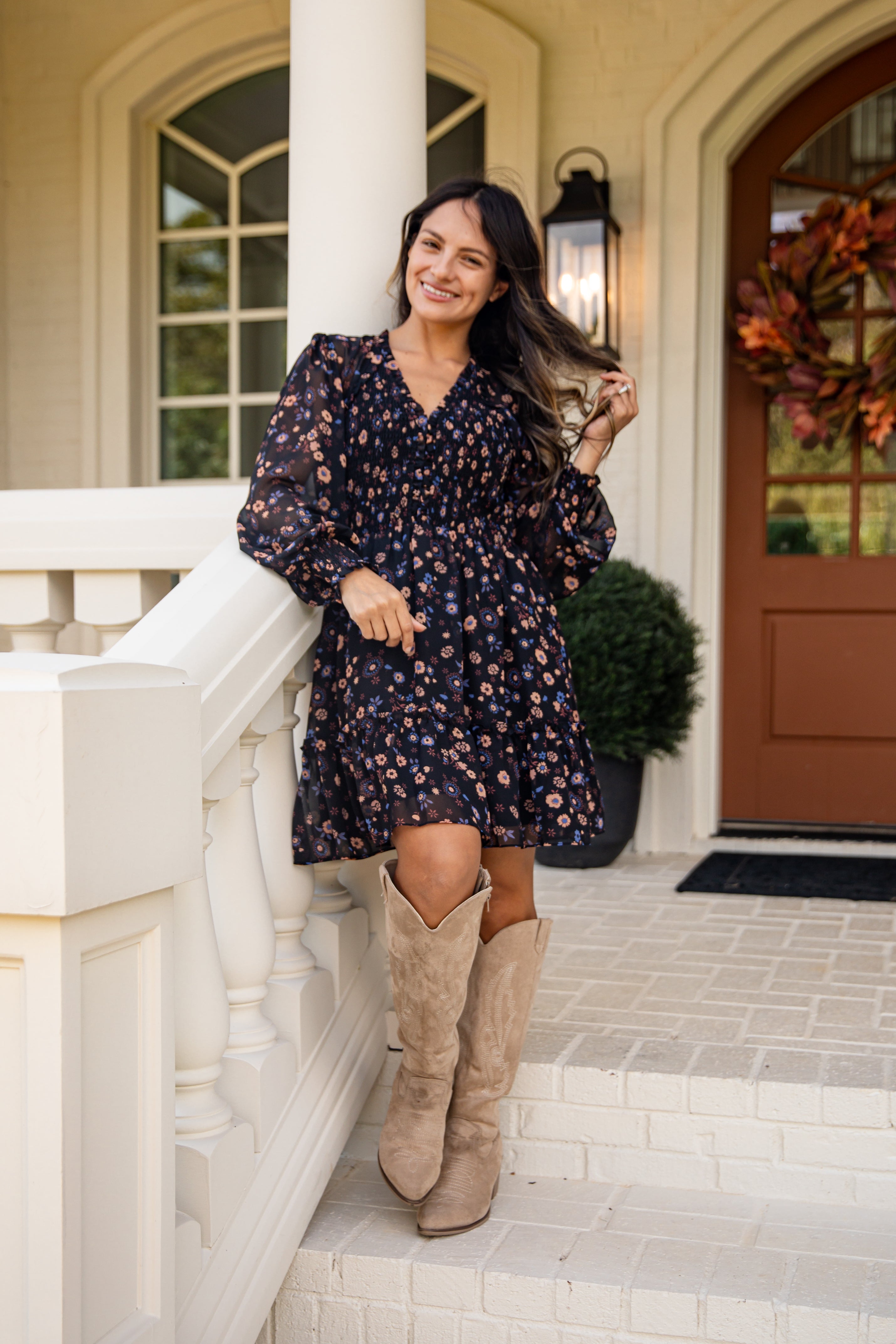 Woman in a floral dress and beige knee-high boots standing on a porch.