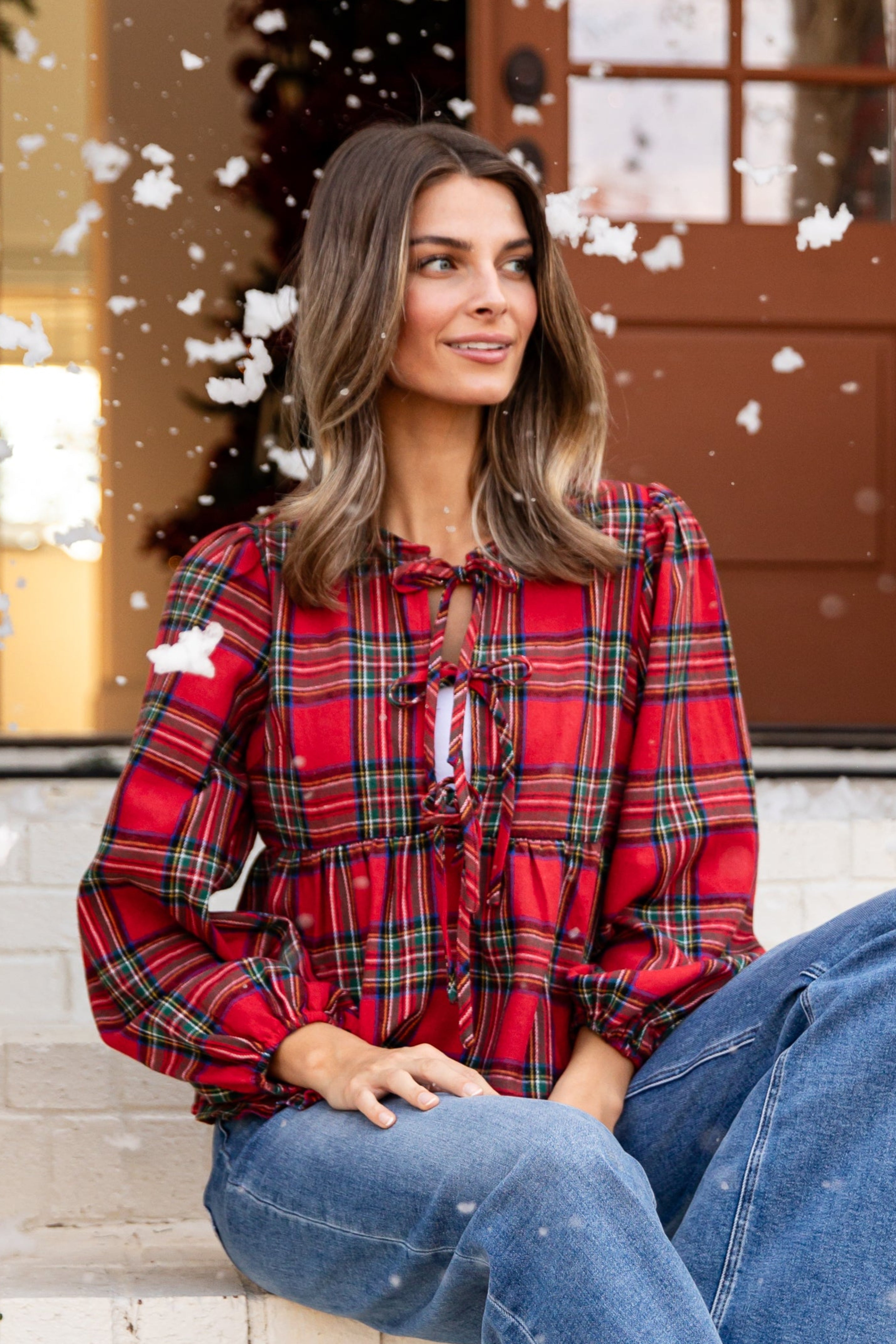 Woman sitting on steps wearing a red plaid shirt and blue jeans, with Christmas decorations in the background.