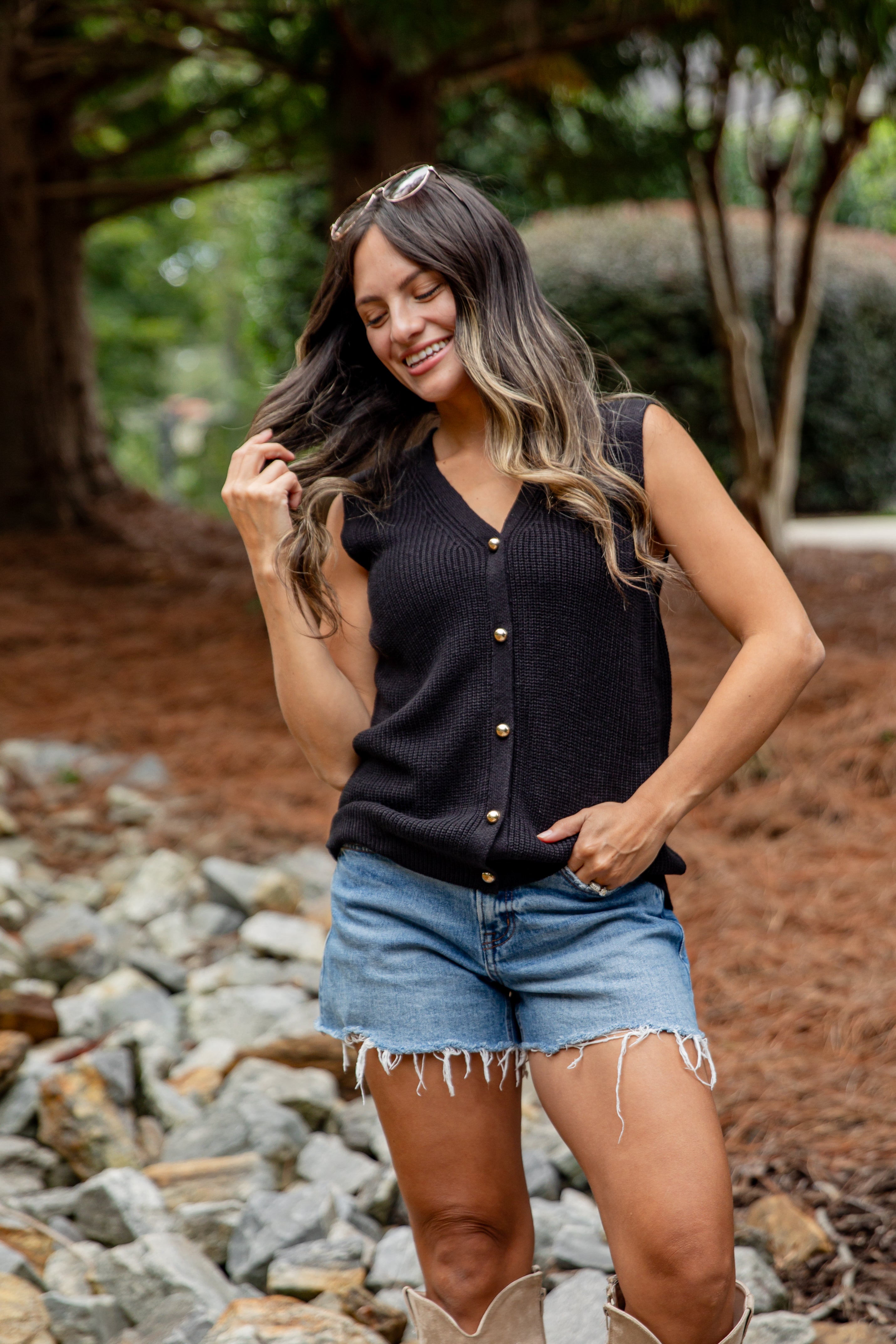 Woman in black sleeveless top and denim shorts standing outdoors with trees and rocks in the background
