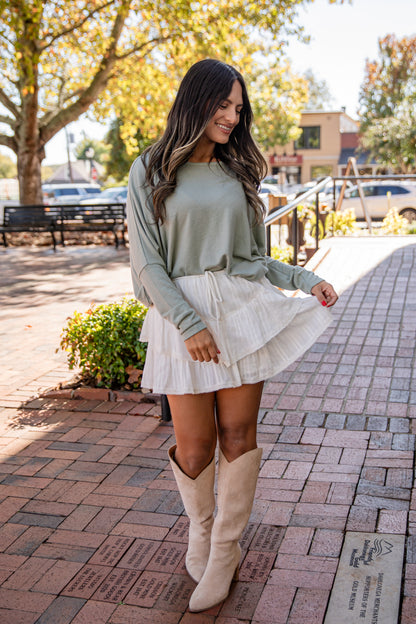 Woman in a light green sweater and white skirt with beige boots walking on a sidewalk.