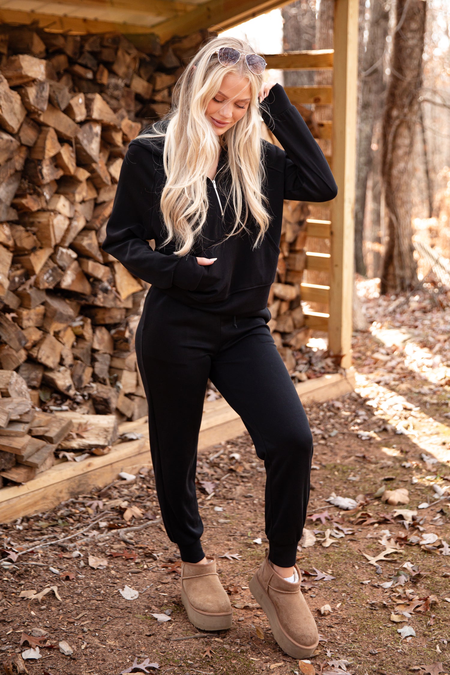 Woman in black outfit standing in front of stacked firewood in a forest setting