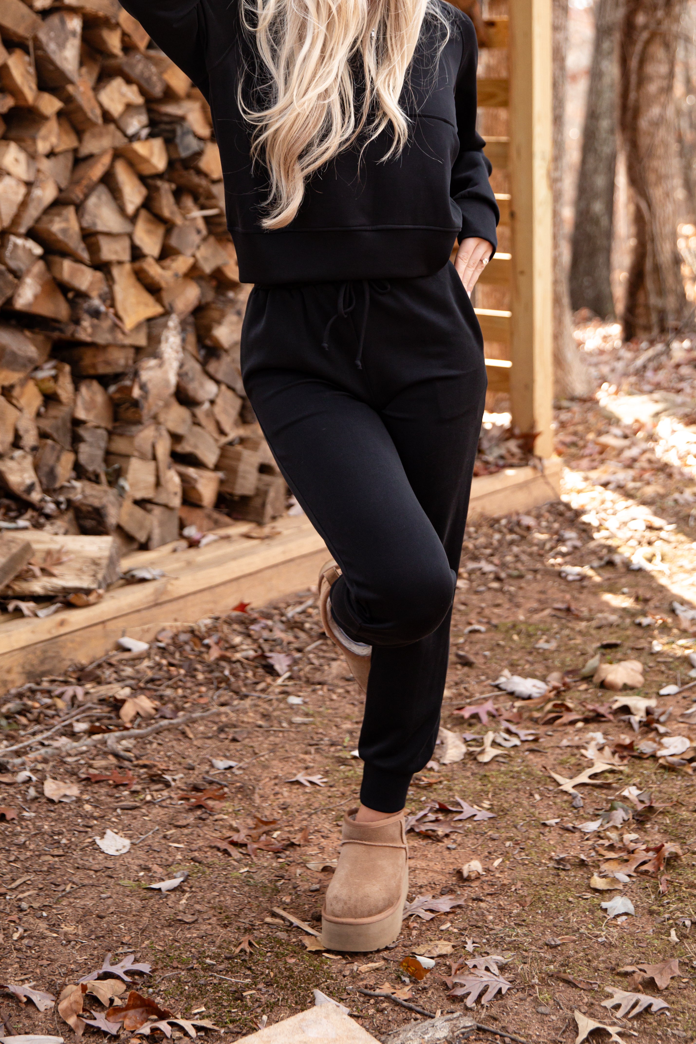 Person wearing a black outfit with beige boots standing in front of stacked firewood.