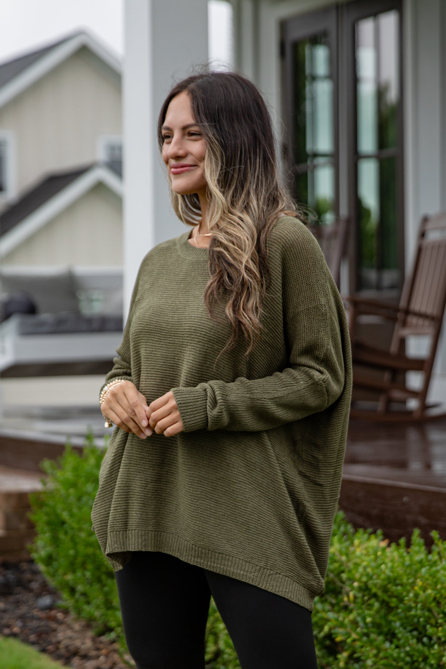 Woman wearing an olive green sweater standing outdoors near a house.