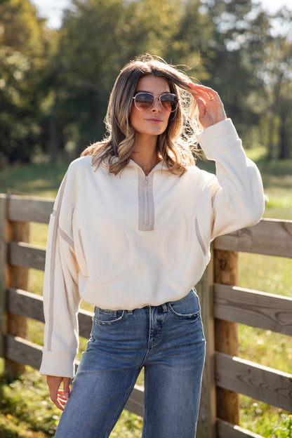 Woman wearing a cream blouse and blue jeans standing in front of a wooden fence with trees in the background.