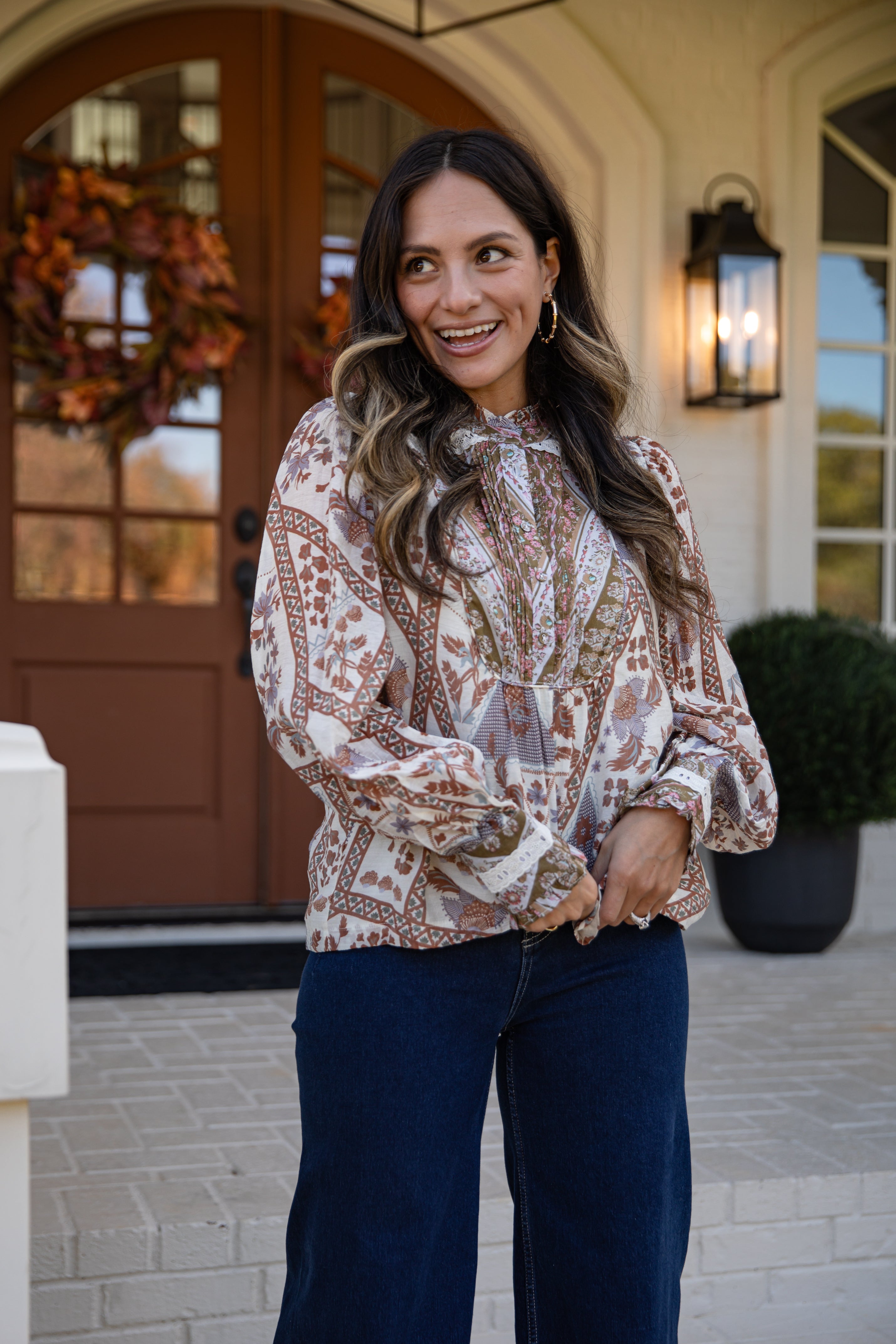 Woman in a patterned blouse and blue pants standing in front of a house entrance.
