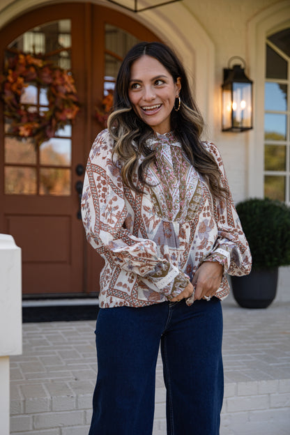 Woman in a patterned blouse and blue pants standing in front of a house entrance.