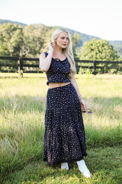 Woman in a black floral dress standing in a grassy field with trees in the background