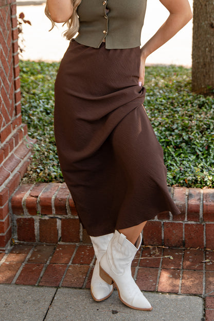 Person wearing a brown skirt and white boots standing on a brick path with greenery in the background