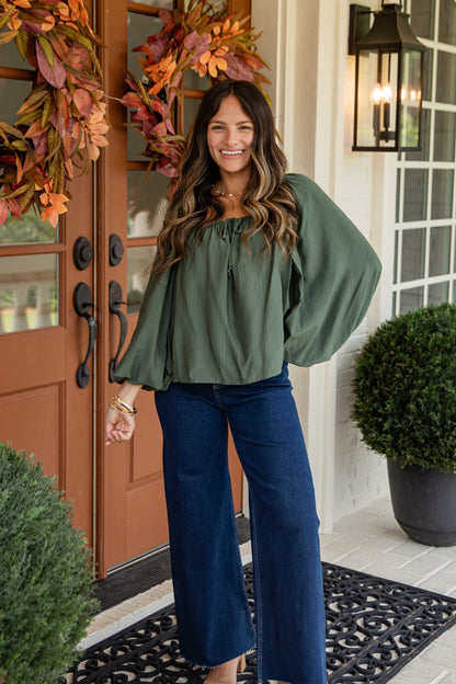 Woman standing in front of a door with autumn wreaths, wearing a green top and blue jeans.
