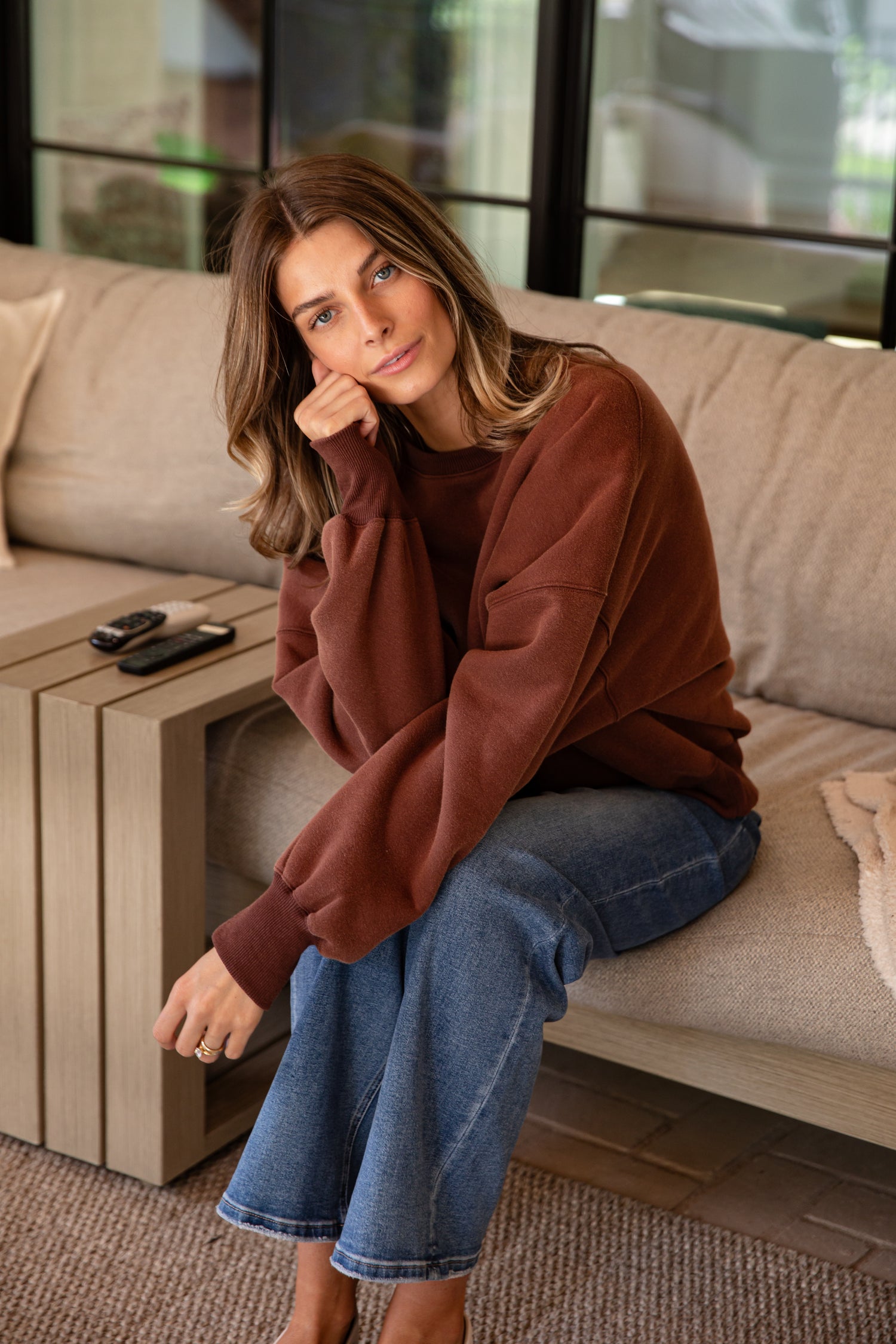 Woman sitting on a couch wearing a brown sweater and blue jeans, with a coffee table and window in the background.