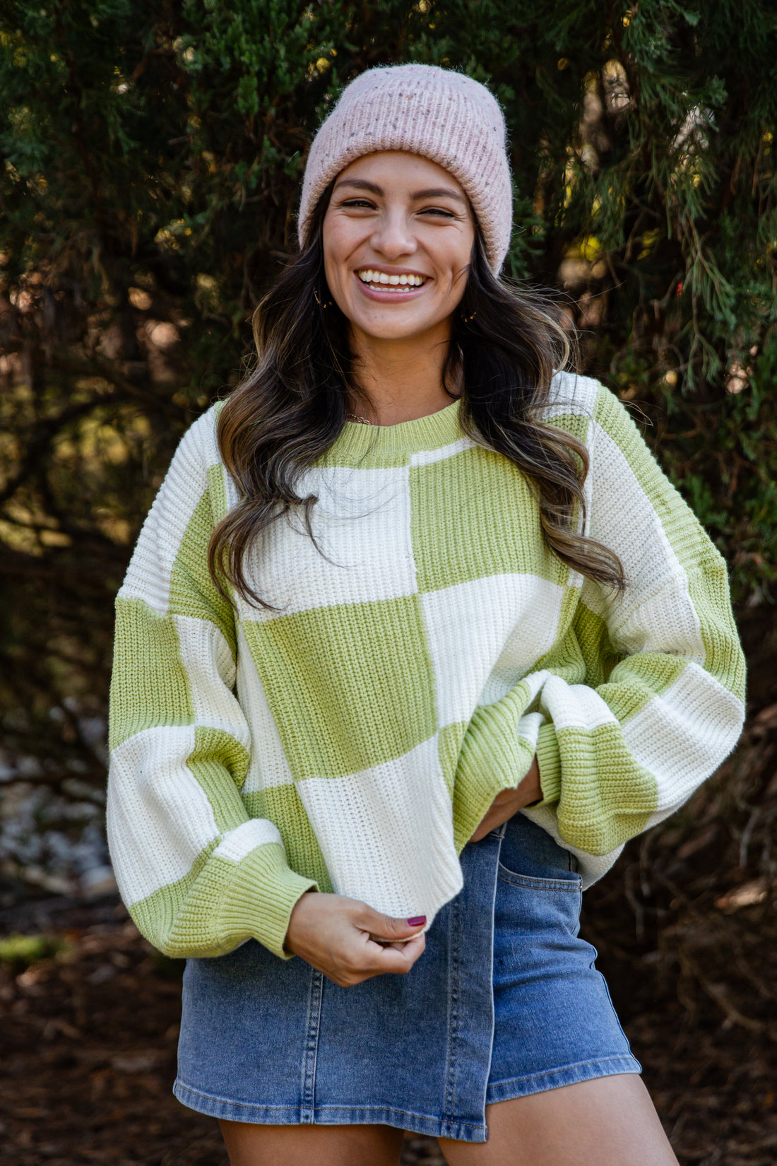Woman wearing a green and white checkered sweater with a pink beanie and denim shorts outdoors.
