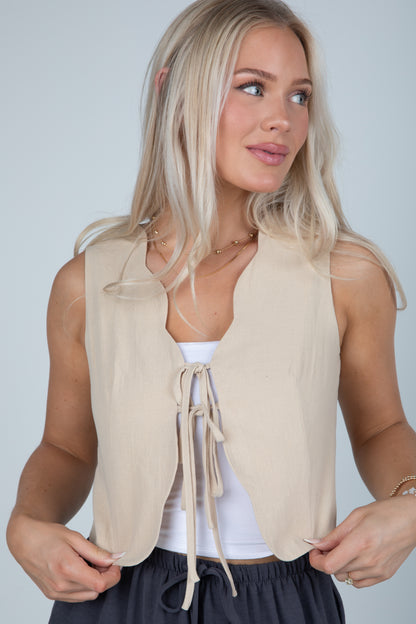 Woman wearing a beige sleeveless top with a white shirt underneath on a plain background