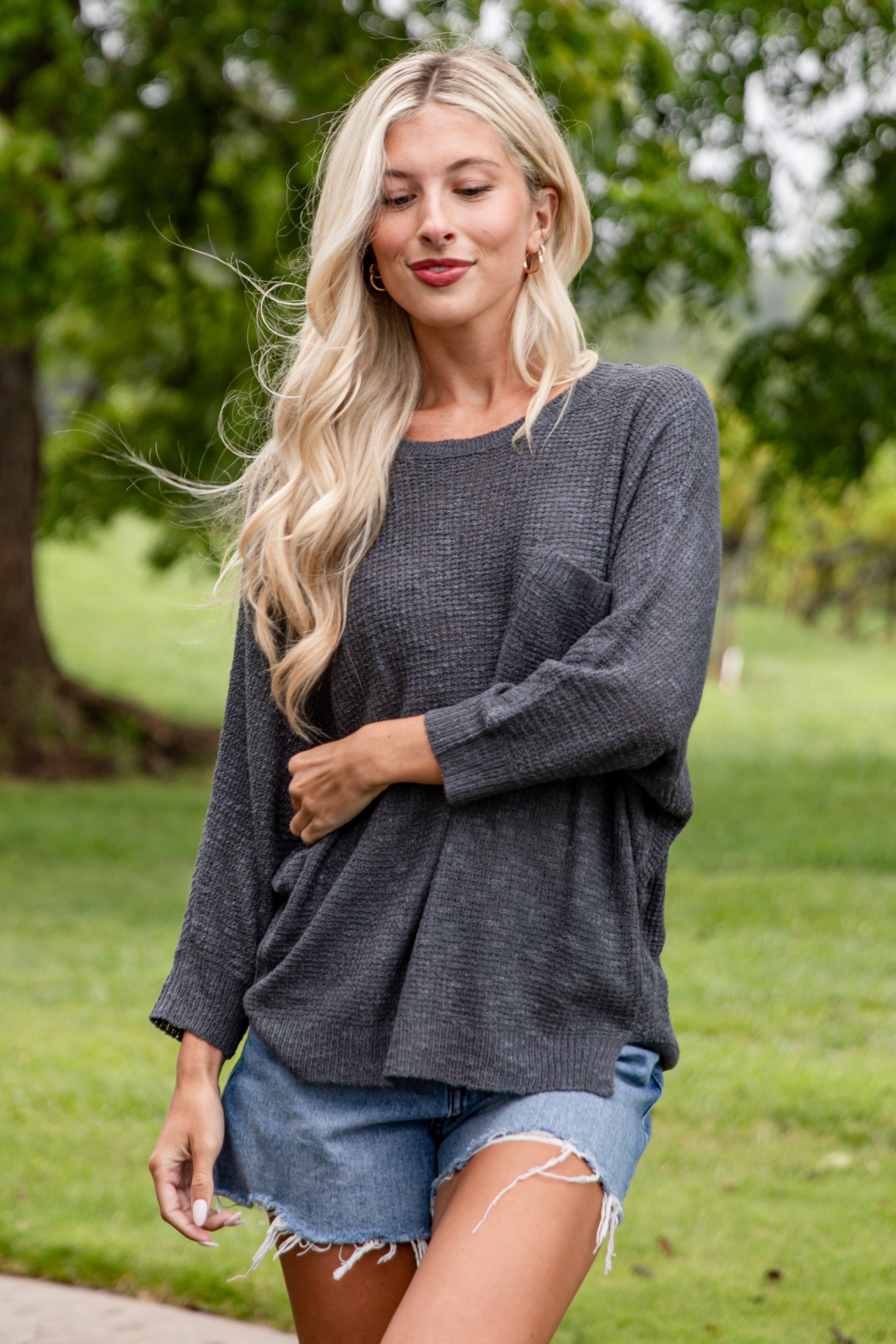 Woman wearing a gray sweater and denim shorts standing outdoors with greenery in the background