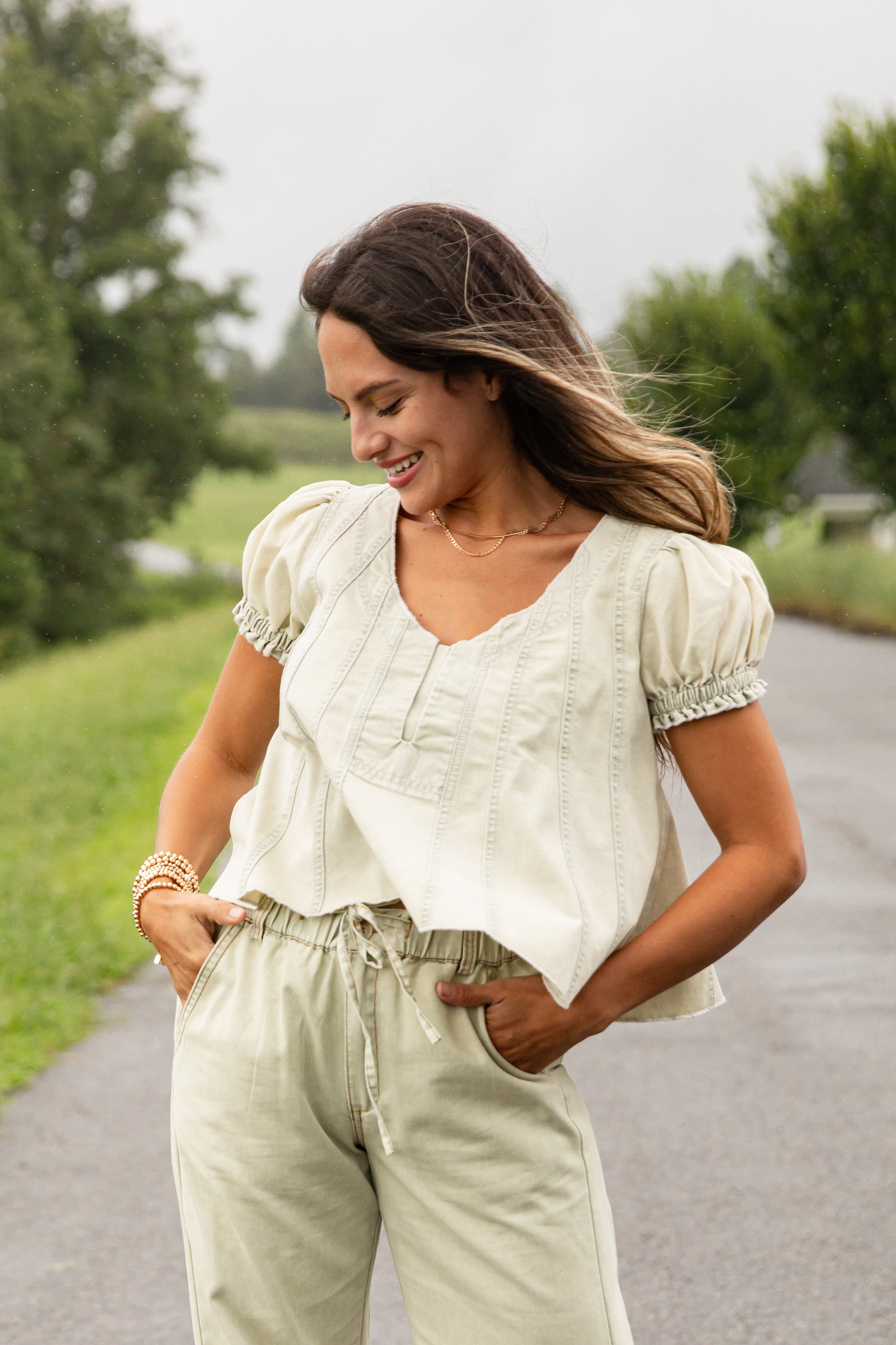 Woman in a white blouse and light green pants standing on a road with greenery in the background