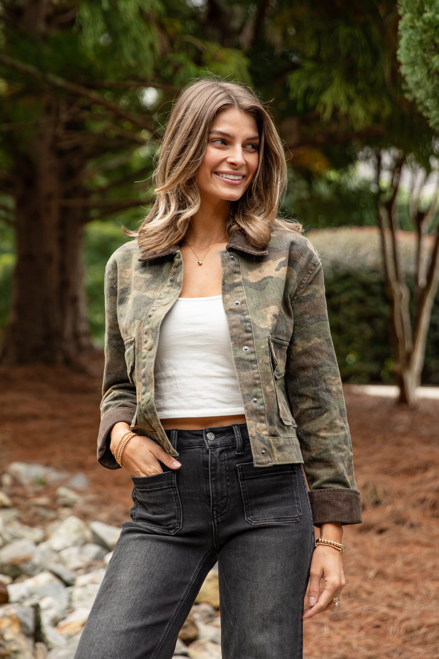 Woman wearing a camouflage jacket, white top, and dark jeans outdoors.
