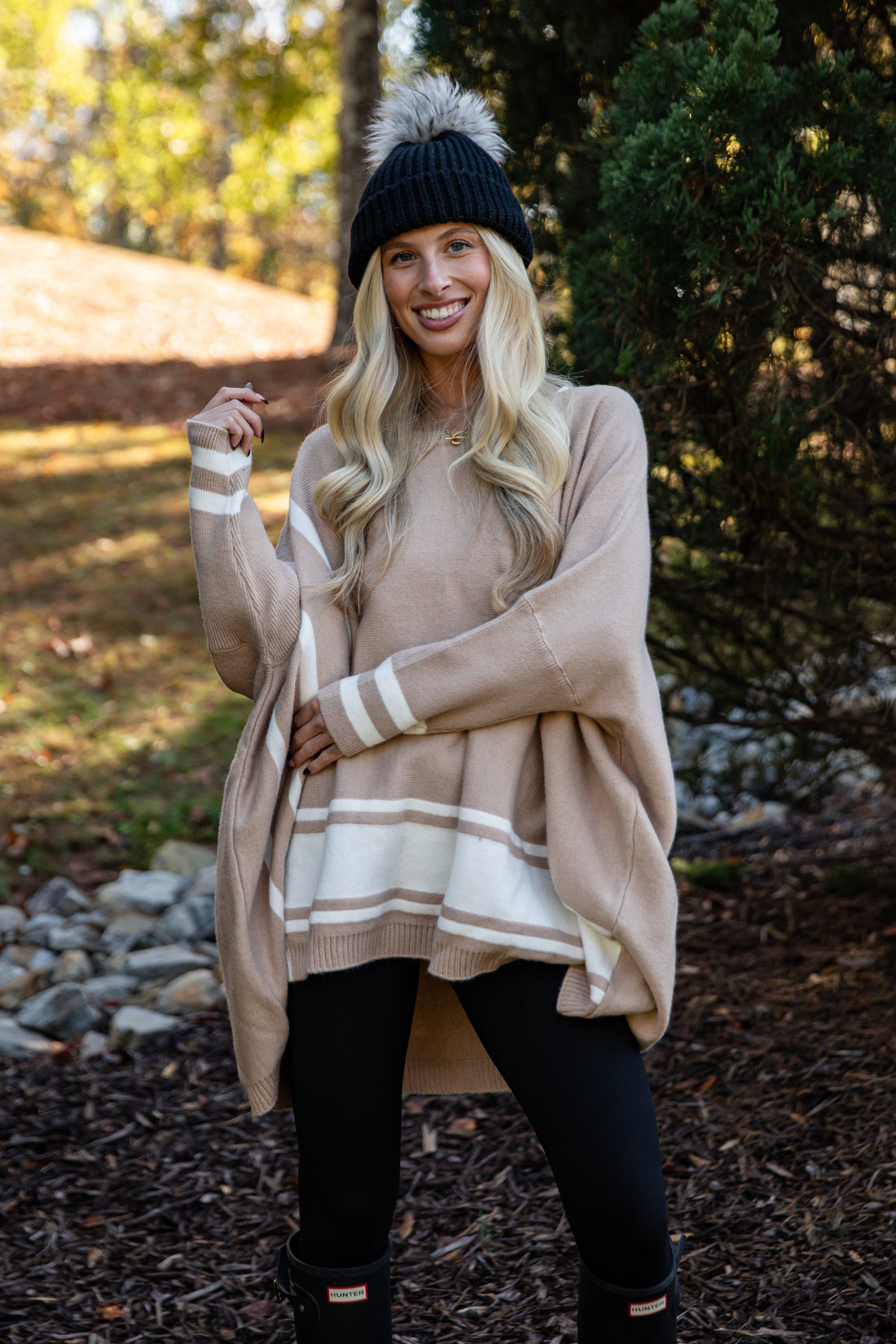 Woman wearing a beige poncho with white stripes, black beanie, and leggings outdoors.