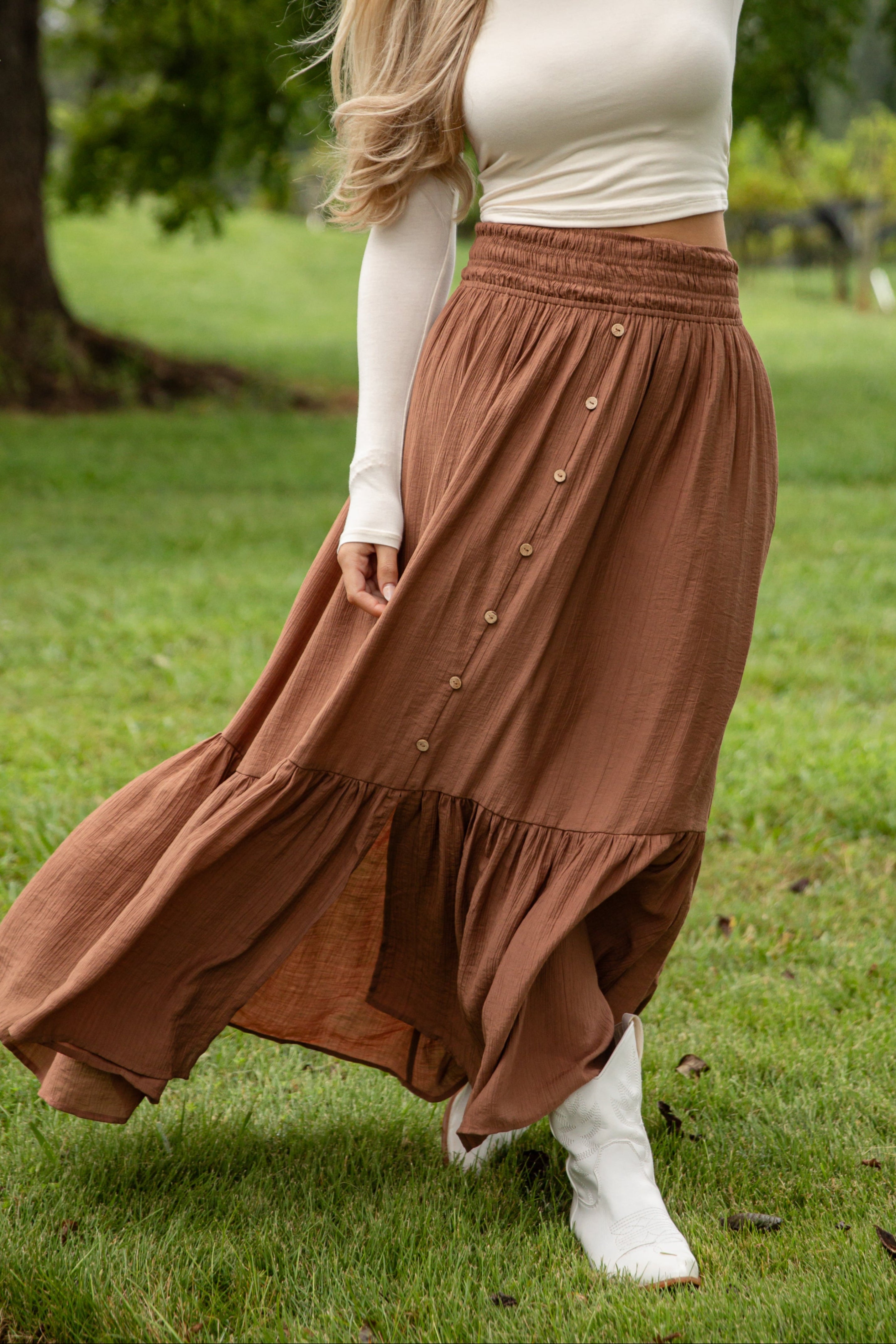 Woman in a white top and brown skirt standing in a grassy outdoor setting.