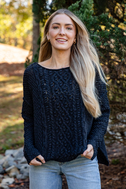 Woman wearing a dark blue knitted sweater outdoors with trees and rocks in the background