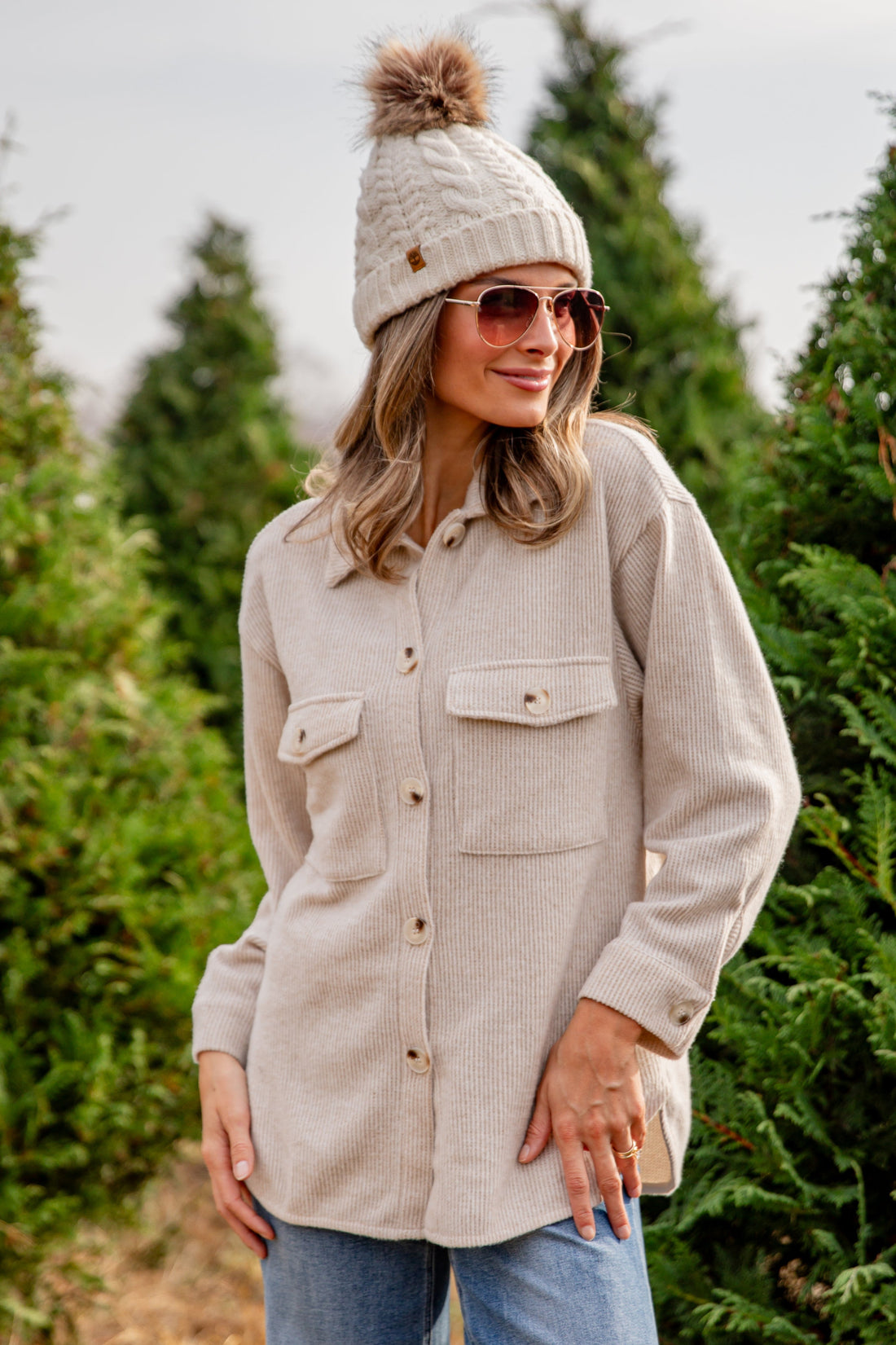 Woman wearing a beige jacket, beanie, and sunglasses standing in a tree farm.