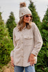Woman wearing a beige jacket, beanie, and sunglasses standing in a tree farm.
