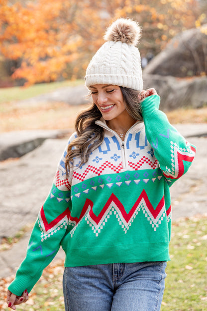 Woman wearing a green sweater with a pattern, standing in a park with autumn foliage.