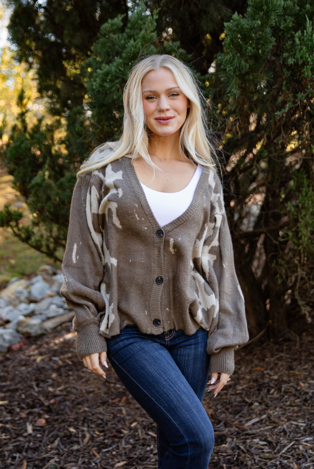 Woman wearing a patterned cardigan and jeans standing outdoors with trees in the background