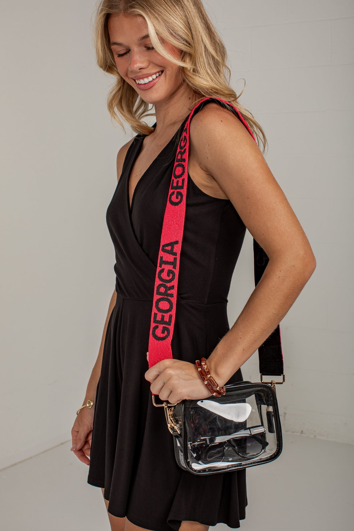 A woman wearing a black dress and holding a clear plastic crossbody bag with a bright pink strap printed with the word &