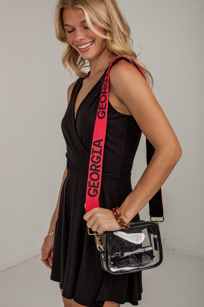 A woman wearing a black dress and holding a clear plastic crossbody bag with a bright pink strap printed with the word &