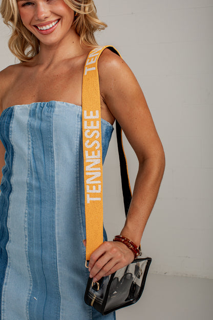 A woman wearing a blue denim strapless dress carries a small black clear handbag with a wide mustard yellow shoulder strap printed with &