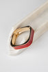 A geometric ring with a gold-toned metal band and a dual-tone red and black enamel inlay, displayed on a white marble stand against a light gray background.
