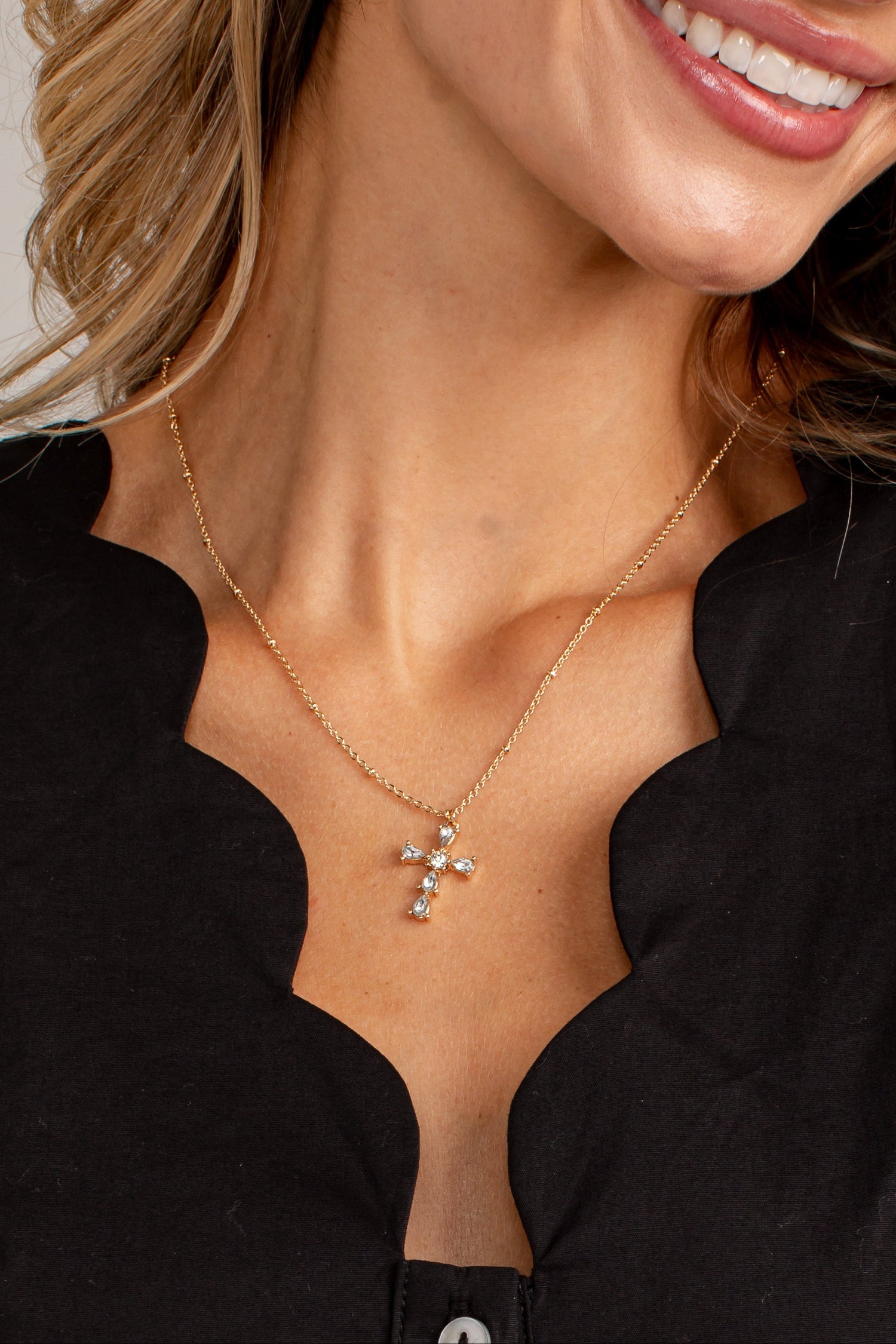 A close-up of a woman wearing a delicate gold cross pendant necklace with small clear stones, styled with a black scalloped top.