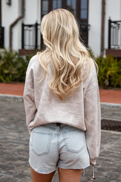 Woman wearing a beige sweatshirt and light denim shorts with long blonde hair, standing outdoors.