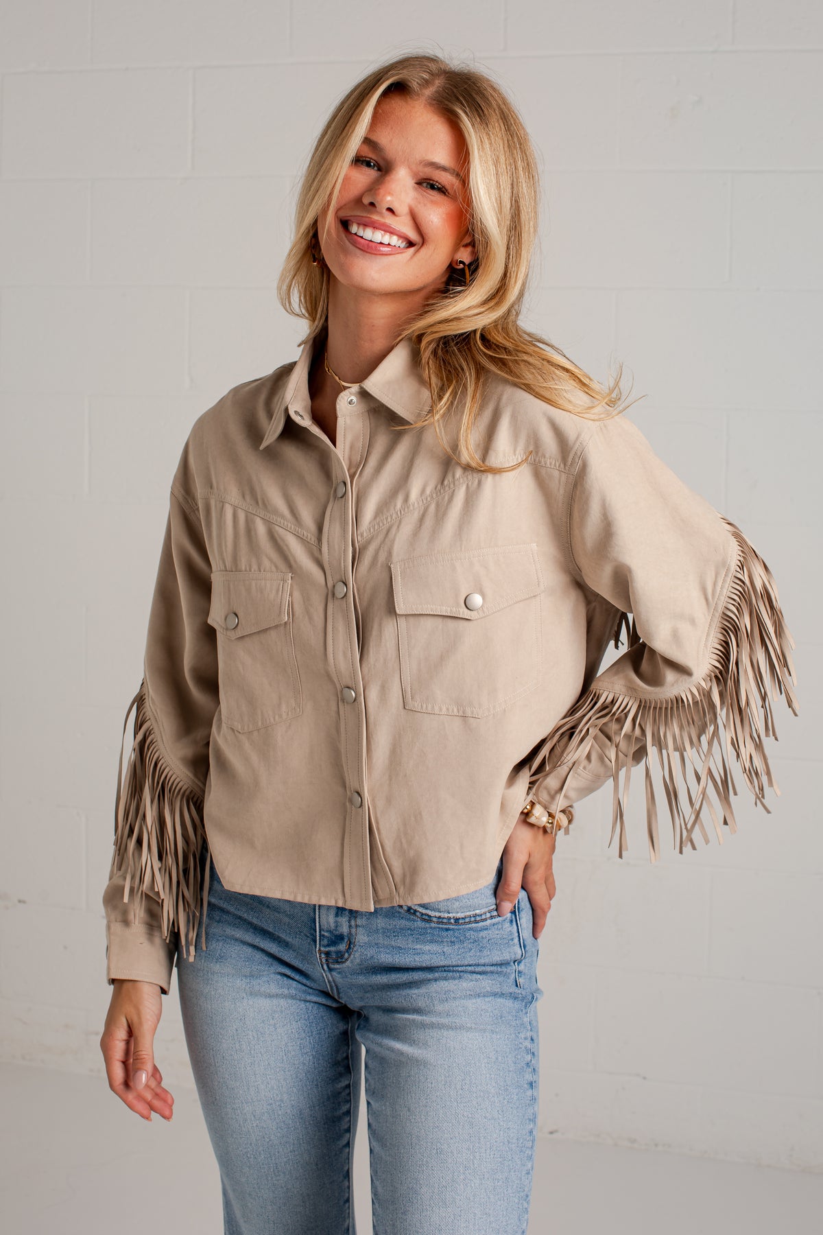 A woman wearing a beige long-sleeve button-up shirt with fringe detailing on the sleeves, paired with blue jeans, standing against a light gray brick wall.