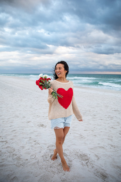 Woman on a beach holding flowers and wearing a sweater with a heart design.