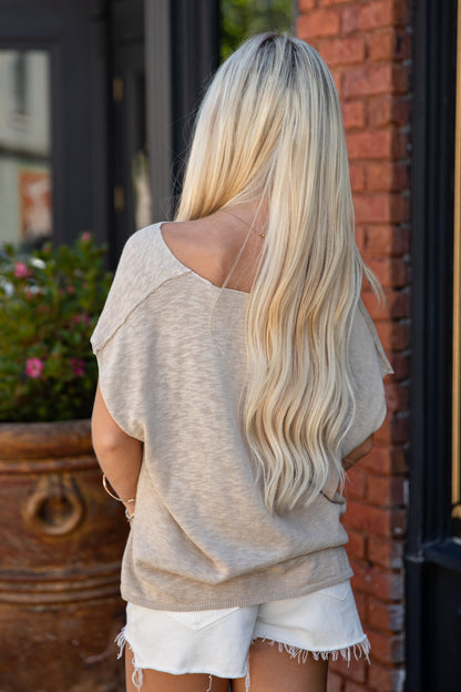 A woman with long blonde hair is shown from behind, wearing a loose beige knit top and white frayed denim shorts, standing outdoors next to a brick wall.