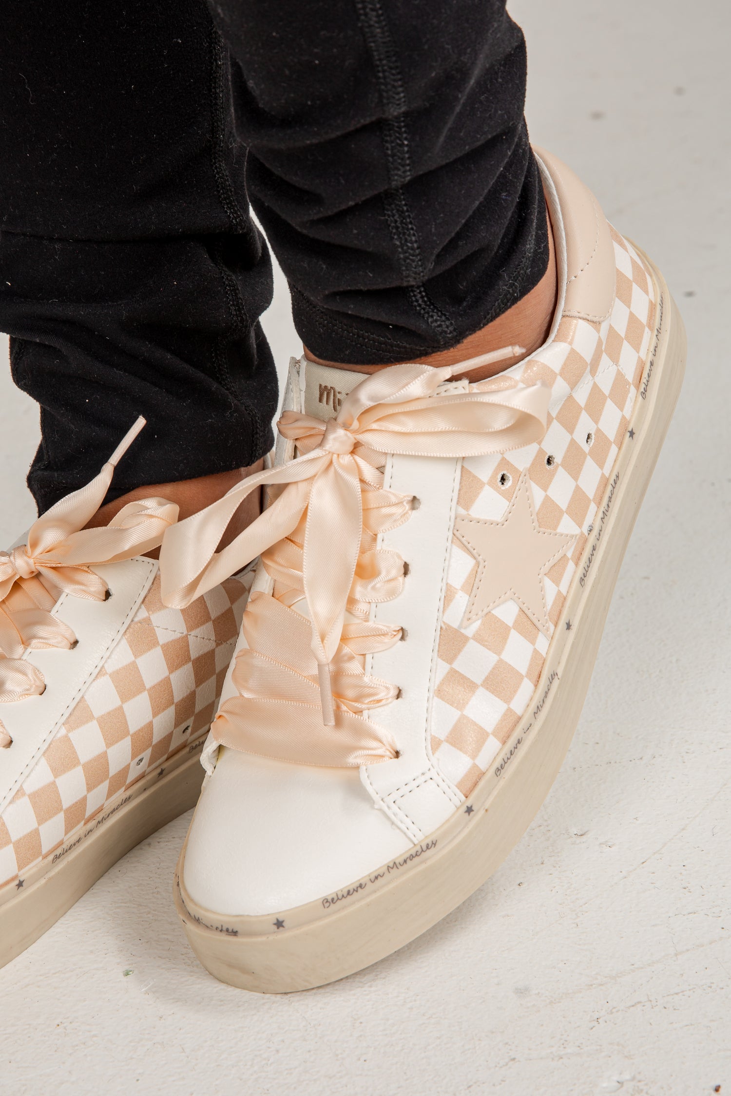 Beige checkered sneakers with ribbons worn by a person on a light gray floor.