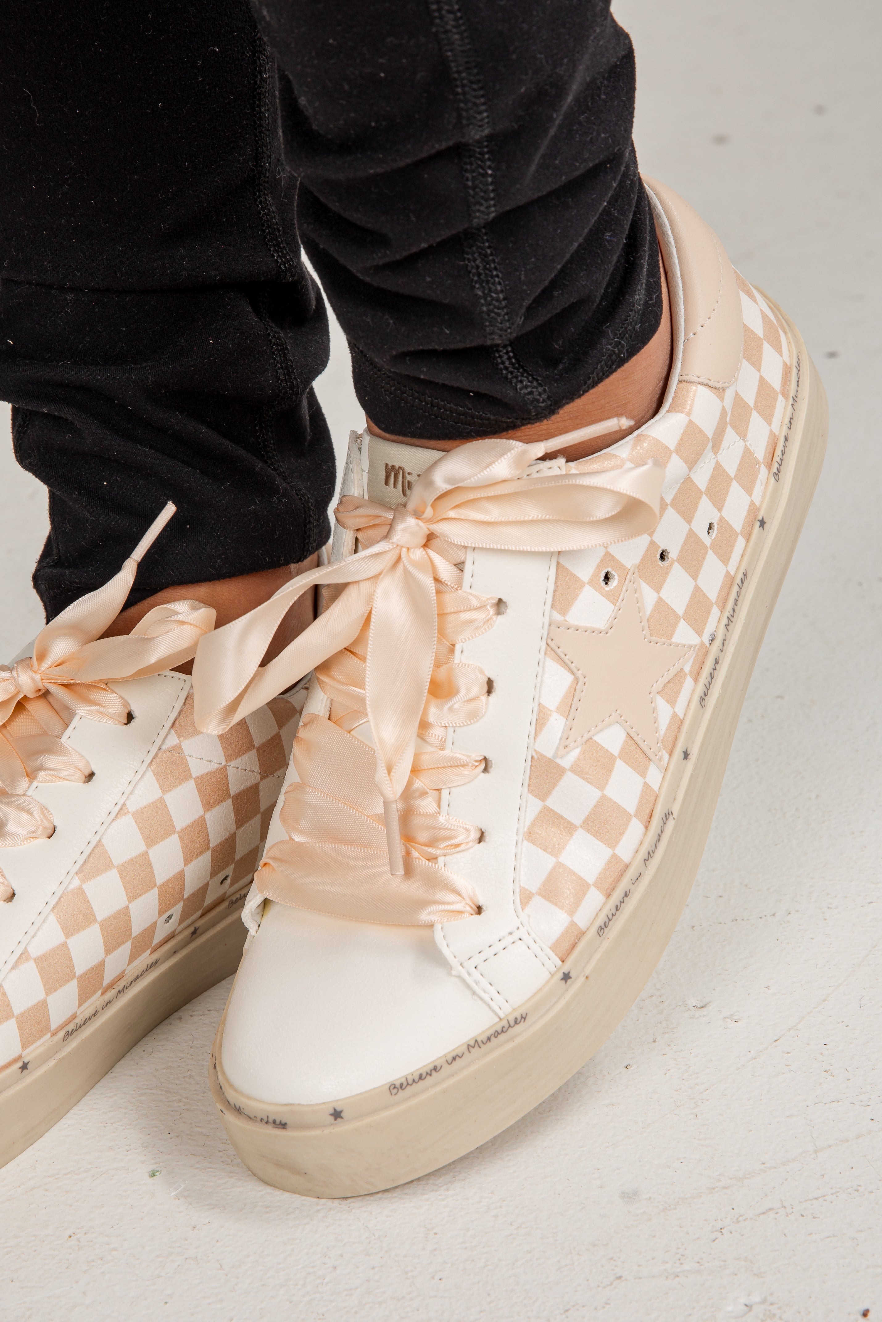 Beige checkered sneakers with ribbons worn by a person on a light gray floor.