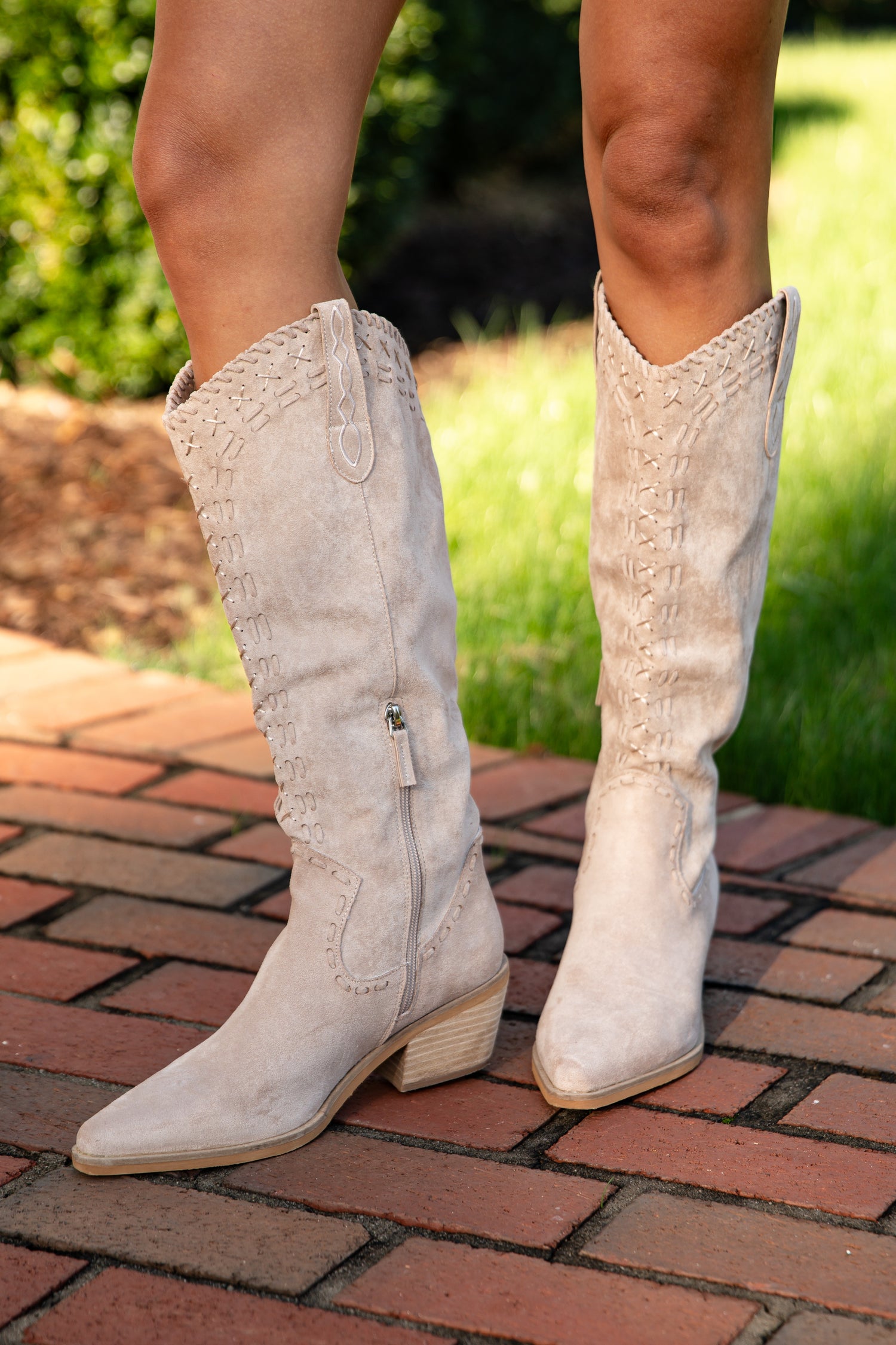 White knee-high boots on a person standing on a brick path with greenery in the background