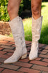 White knee-high boots on a person standing on a brick path with greenery in the background