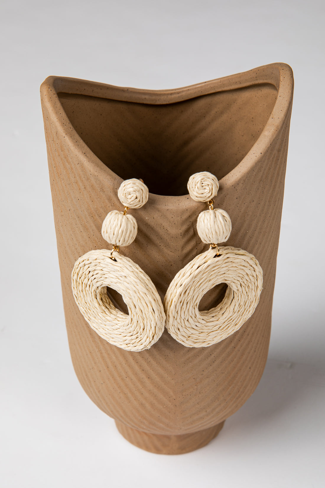 Pair of beige raffia earrings displayed on a textured brown vase.