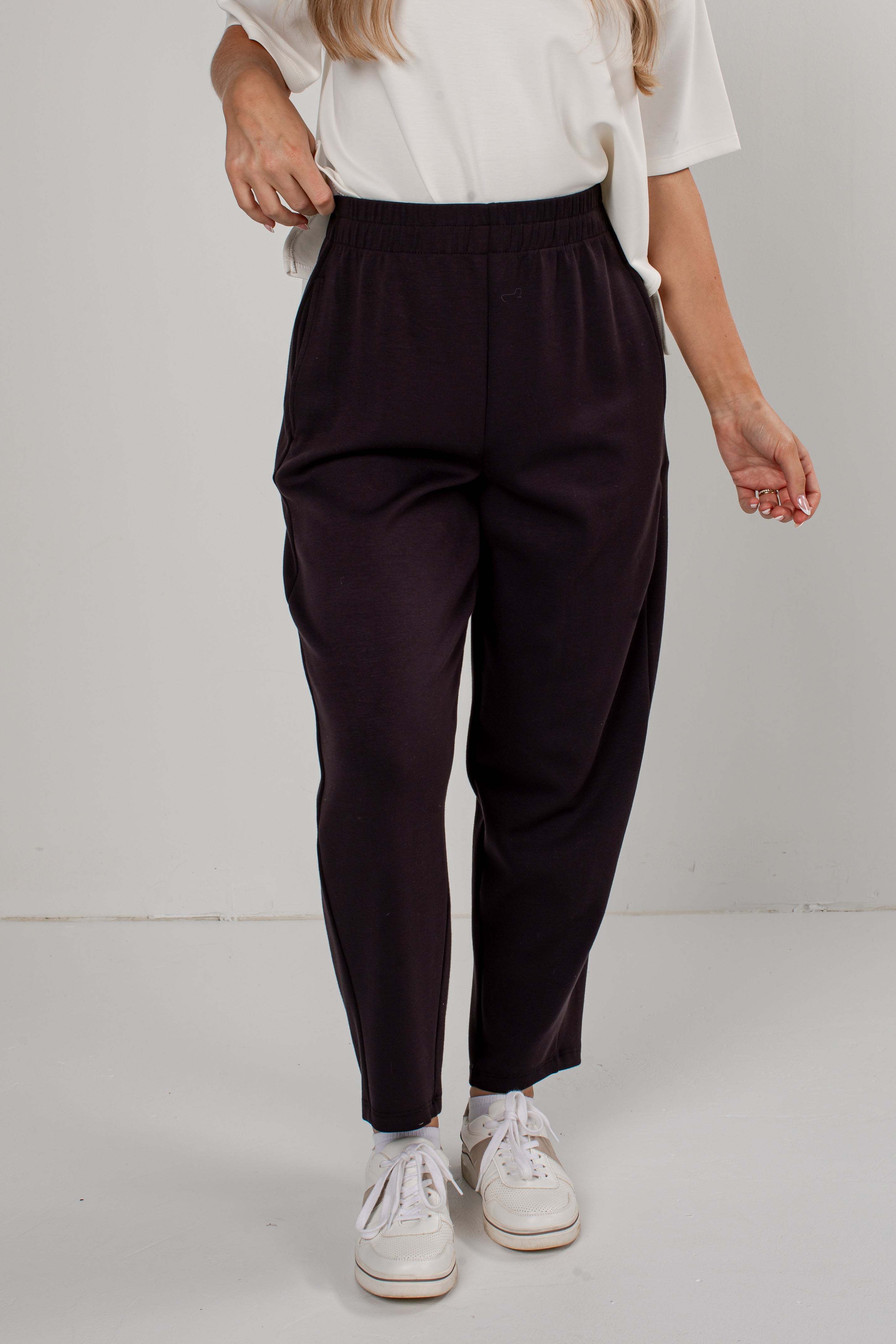 A person wearing dark black tapered pants with an elastic waistband, styled with a white top and white sneakers against a plain light gray studio background.