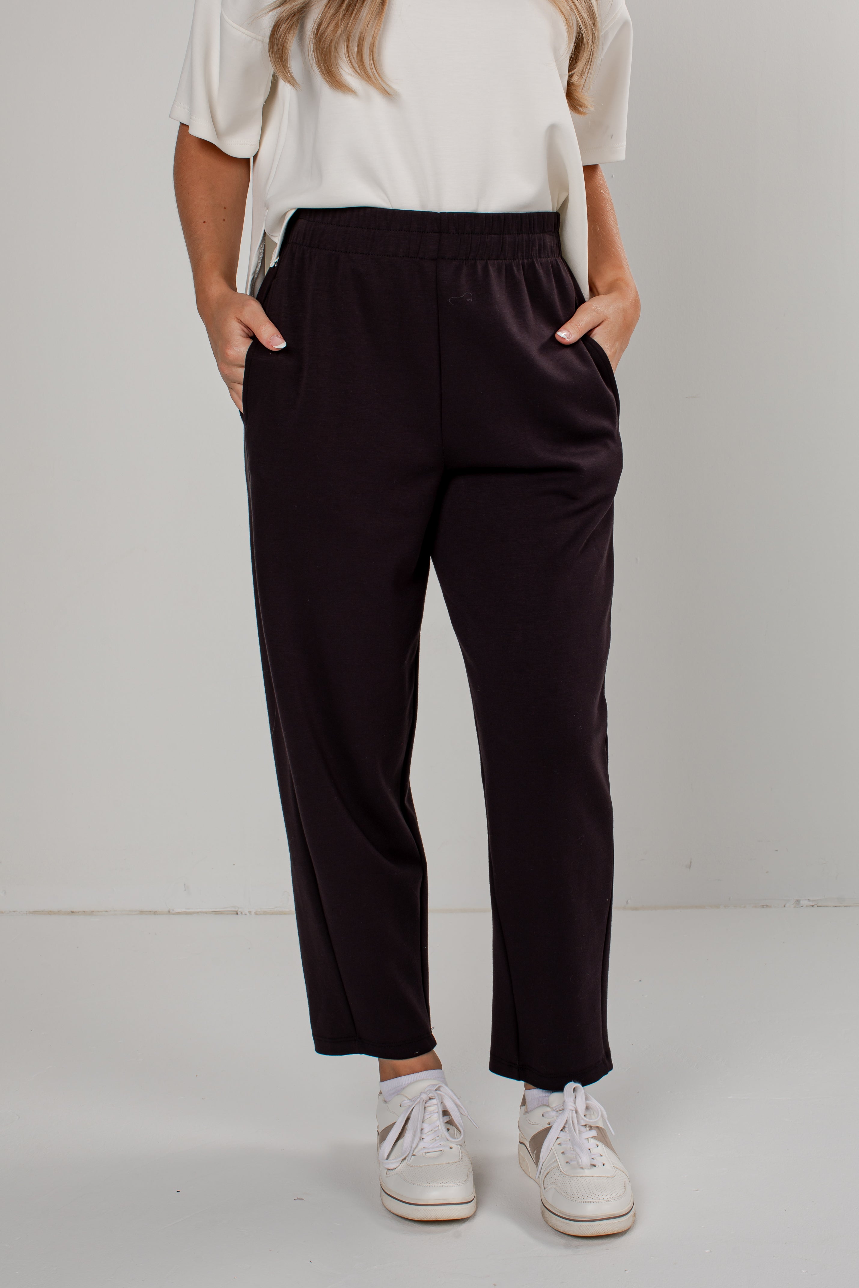 A person wearing dark plum-colored wide-leg cropped pants with an elastic waistband and side pockets, paired with a white top and white sneakers, standing against a light gray studio background.