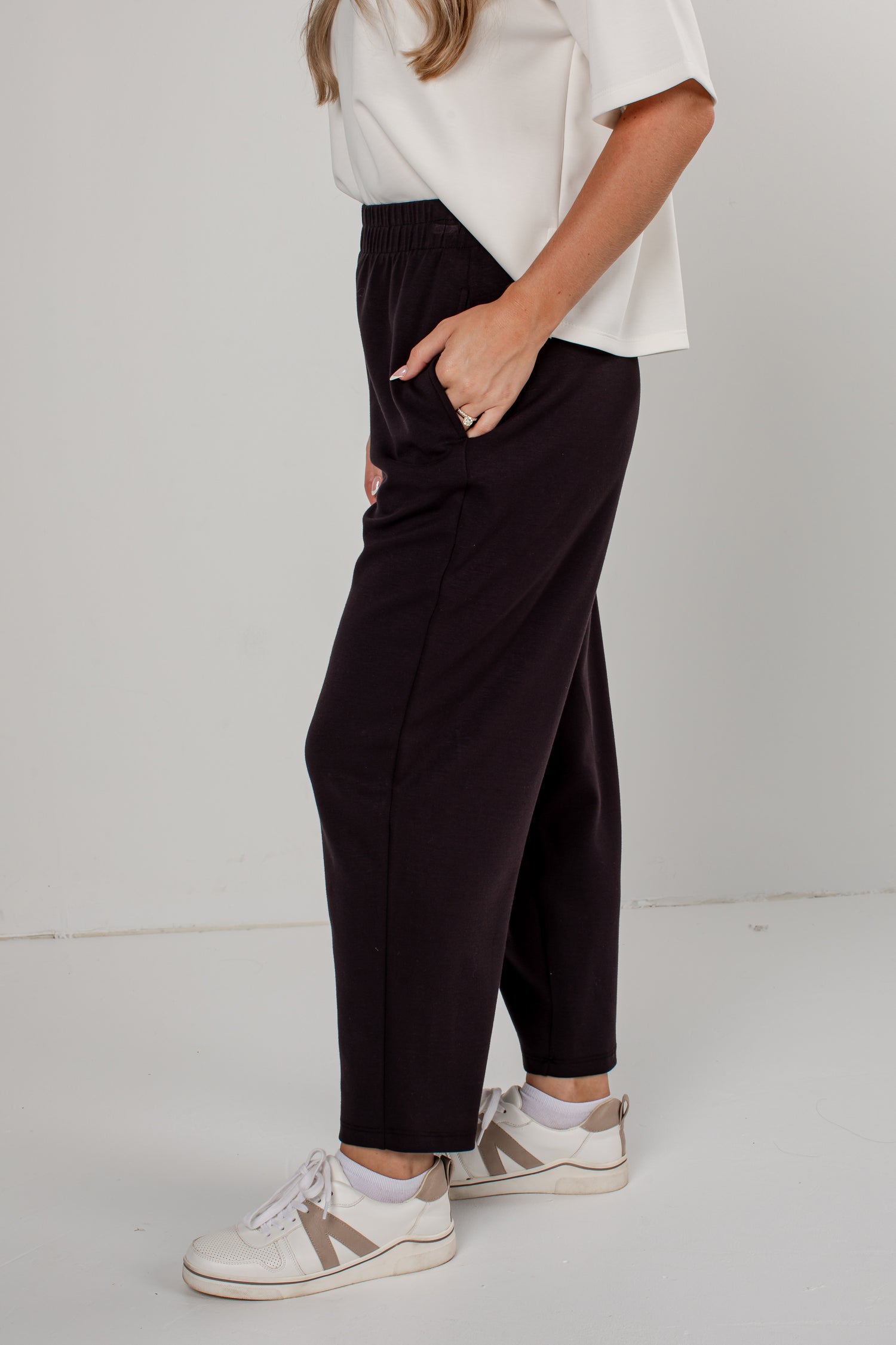 A person wearing dark black-colored wide-leg cropped pants, a white short-sleeve top, and white sneakers with beige stripes, standing against a light gray studio background.