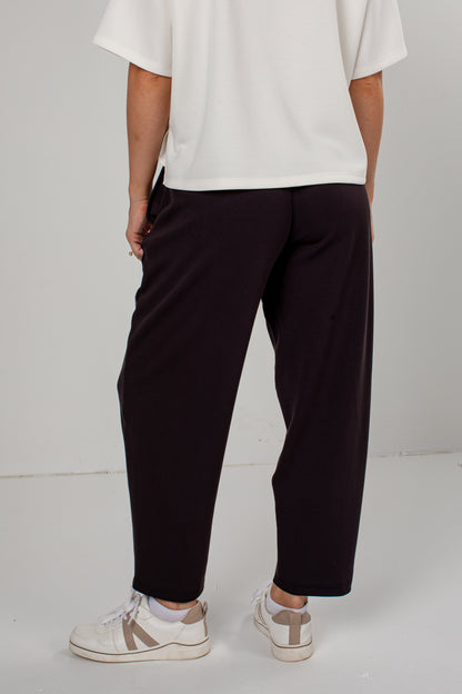 A person shown from the back wearing dark black-colored wide-leg cropped pants, a white t-shirt, and white sneakers with beige accents, set against a plain light gray studio background.