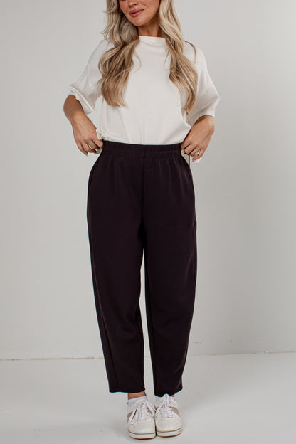 A woman modeling a pair of dark plum-colored wide-leg cropped pants, paired with a white short-sleeve top and white sneakers, against a plain light gray studio background.