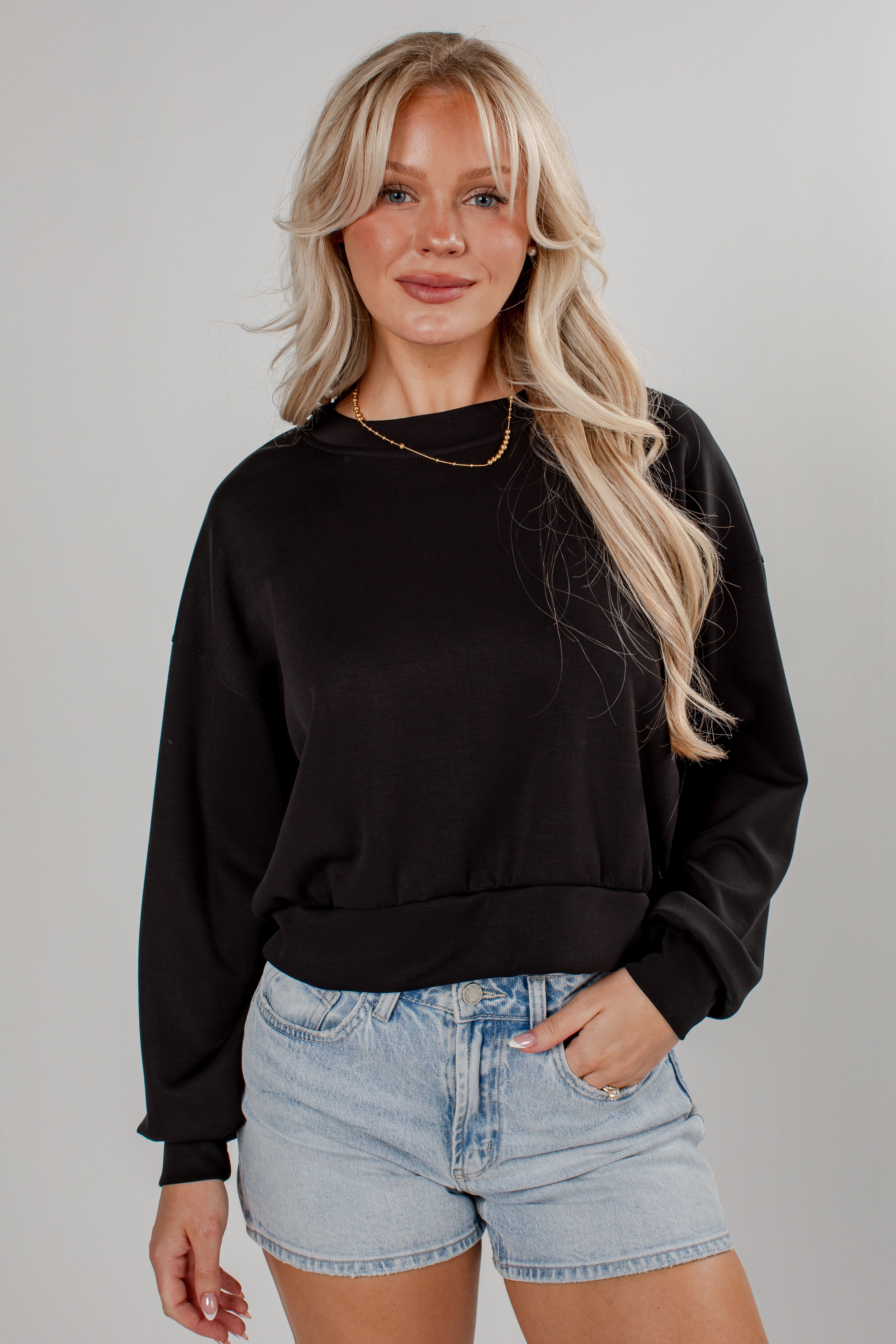 A blonde woman wearing a black long-sleeve crewneck sweatshirt and light-wash denim shorts, standing against a neutral gray studio background.
