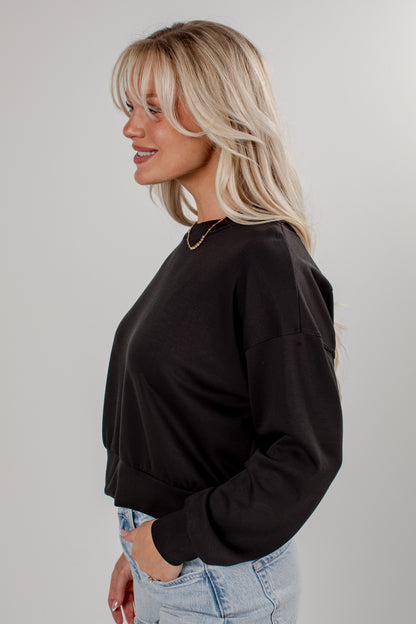 A woman with blonde hair is shown in profile wearing a black long-sleeve crewneck sweatshirt and light blue jeans against a plain light gray studio background.