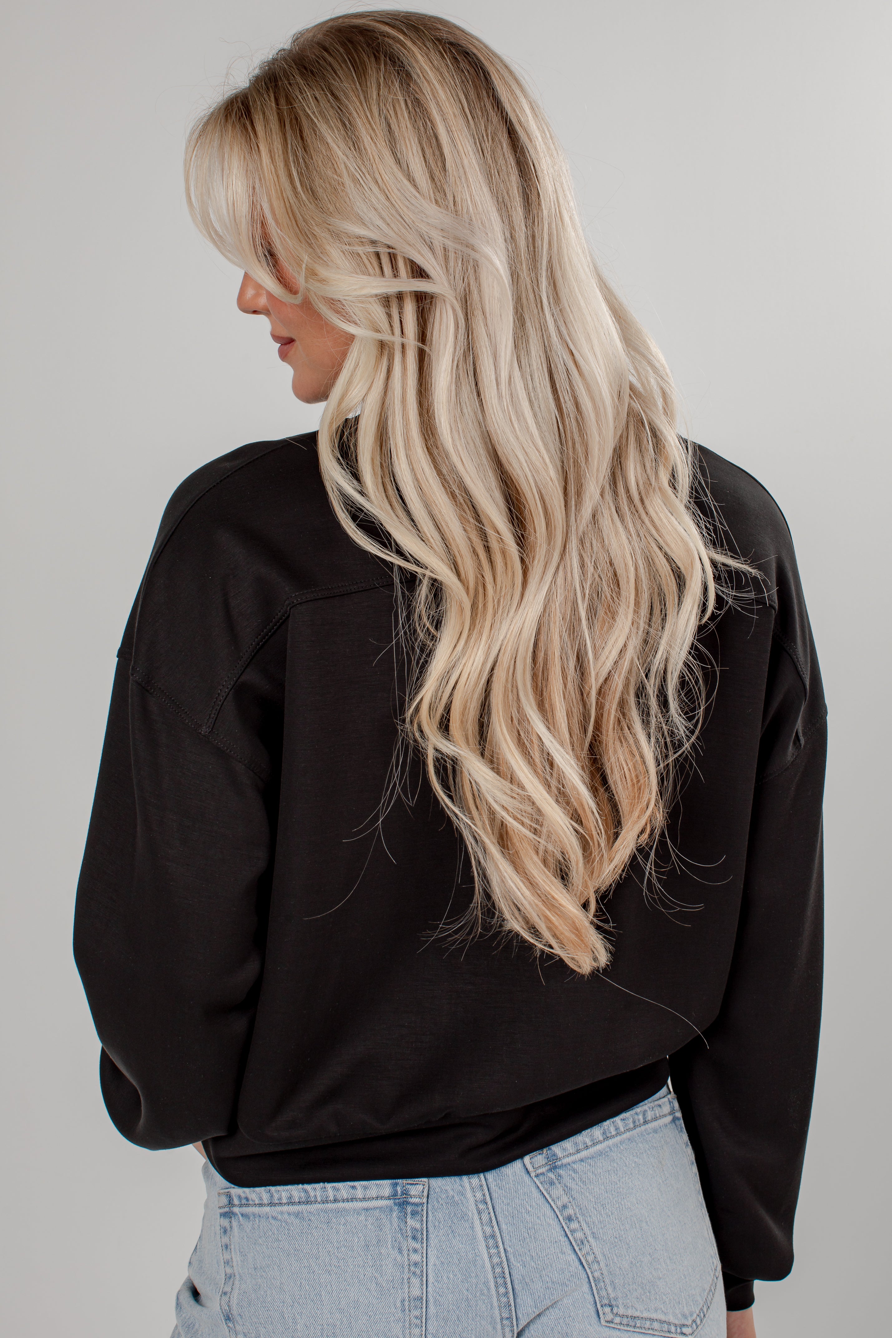 A woman with long, wavy blonde hair is shown from the back, wearing a black long-sleeve top and light blue jeans against a plain light gray background.